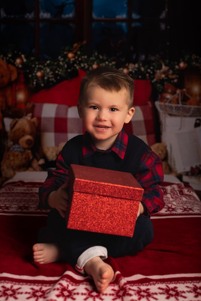 portrait de Noel avec un cadeau- séance photo de Noel à Morillon Petit garçon souriant tenant un cadeau rouge dans un décor de Noel chaleureux