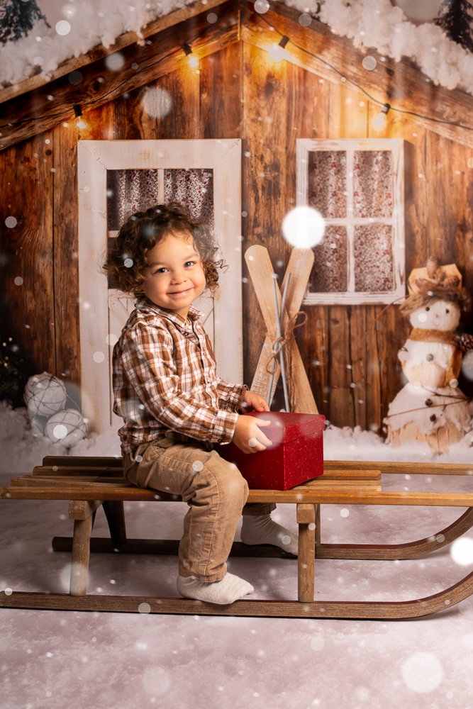 un doux Noêl enneigé-séance photo enfant à Morillon Enfant souriant assis sir un traineau, un cadeau rouge sur les genoux devant un chalet de Noel